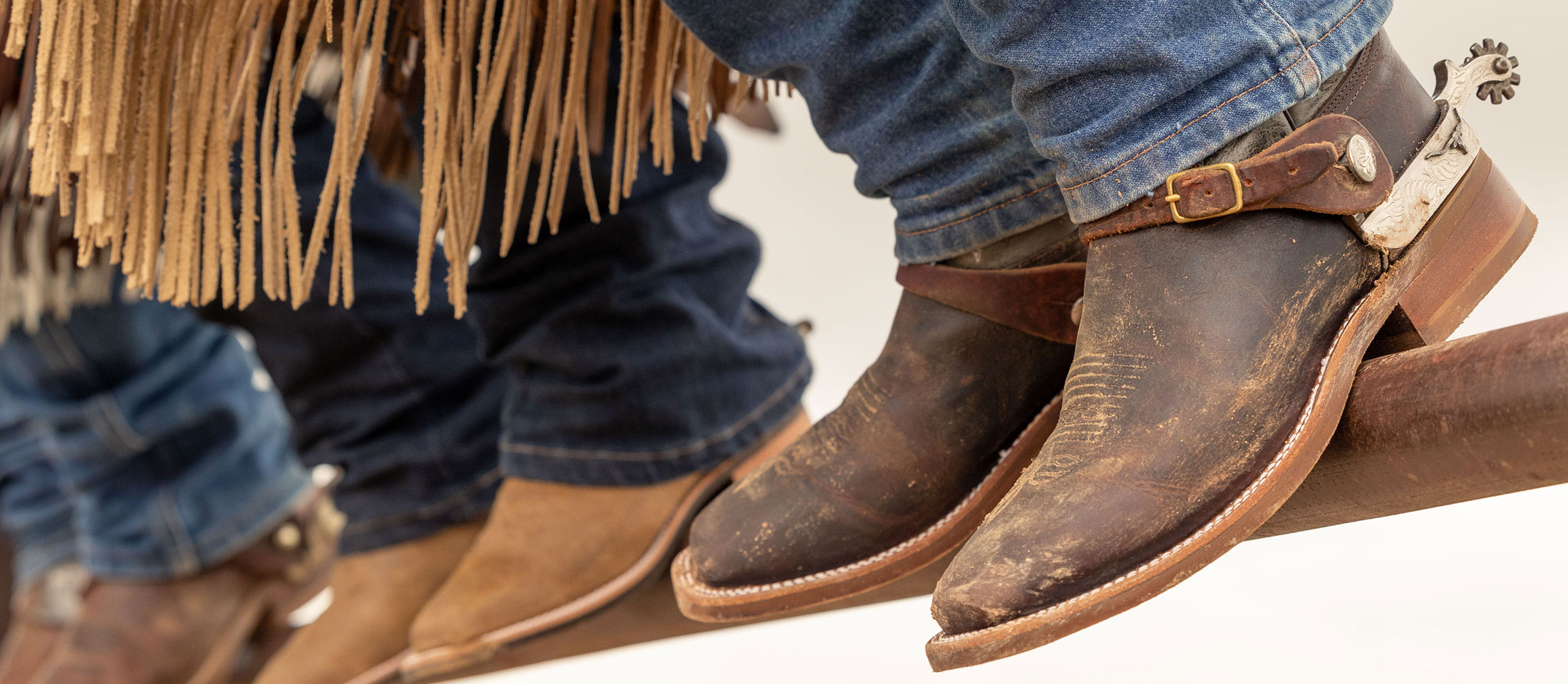 Close up photo of three people sitting on a gate wearing cowboy boots.